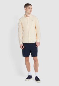 Gestreepte gele longsleeve-shirt met knoopsluiting, gecombineerd met marineblauwe shorts. Het model draagt marineblauwe sneakers en witte sokken. Eenvoudig ontwerp.