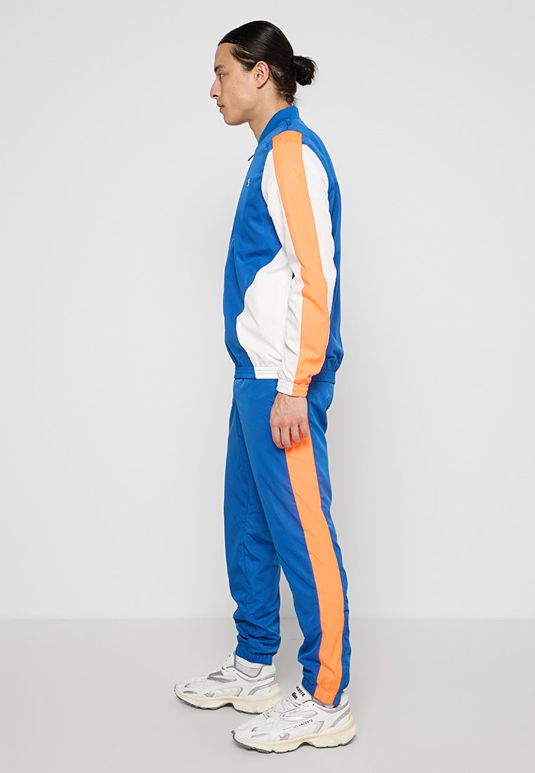 Chándal azul con rayas blancas y naranjas, que incluye una chaqueta con cremallera y pantalones con cintura elástica. Textura suave con un diseño deportivo.