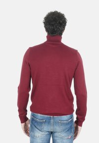 Maglione dolcevita bordeaux, design aderente, collo e polsini a coste, tessuto lavorato a maglia morbido, texture liscia, indossato con jeans di denim blu.