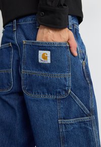 Denim pants with a dark blue finish, featuring multiple pockets and a visible white label with a yellow logo on the back pocket.