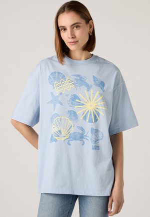 Kvinde iført en lyseblå oversized t-shirt med blå og gule tryk af havdyr og skaller, kombineret med blå jeans.