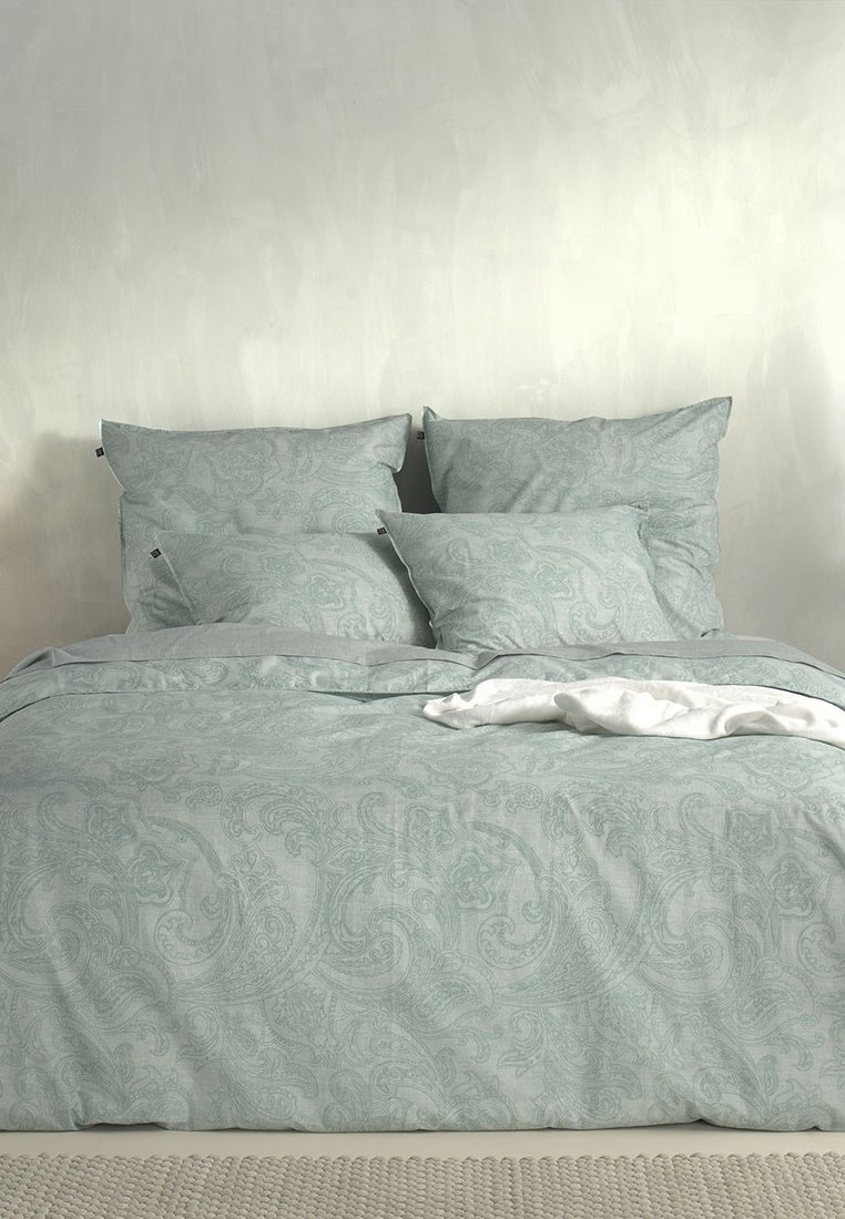 Juego de cama azul pálido con intrincado patrón de paisley, que incluye cuatro almohadas y una suave manta blanca arrugada sobre el edredón.