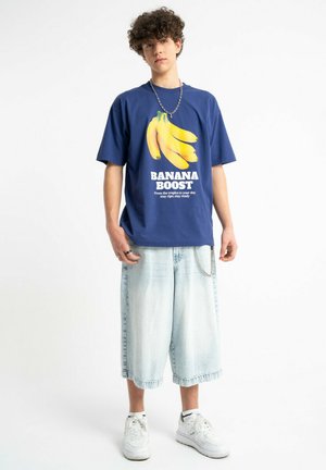 Nastolatek noszący niebieski T-shirt "Banana Boost", jasne długie krótkie spodenki z denimu, białe trampki, łańcuszek na szyi i pierścionki, stojący na białym tle.