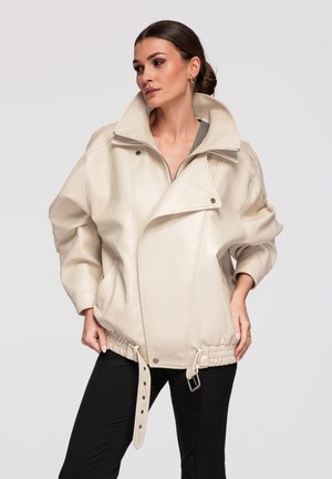 Femme aux cheveux foncés coiffés en chignon bas, portant une grande veste en cuir beige avec ceinture bouclée et pantalon noir, debout devant un fond uni.