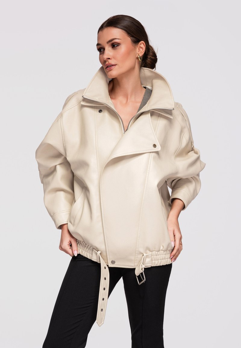 Femme aux cheveux foncés coiffés en chignon bas, portant une grande veste en cuir beige avec ceinture bouclée et pantalon noir, debout devant un fond uni.