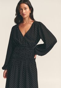 Black dress with white polka dots, deep V-neck, long puff sleeves, and smocked waist detail. Soft fabric with a flowing silhouette.