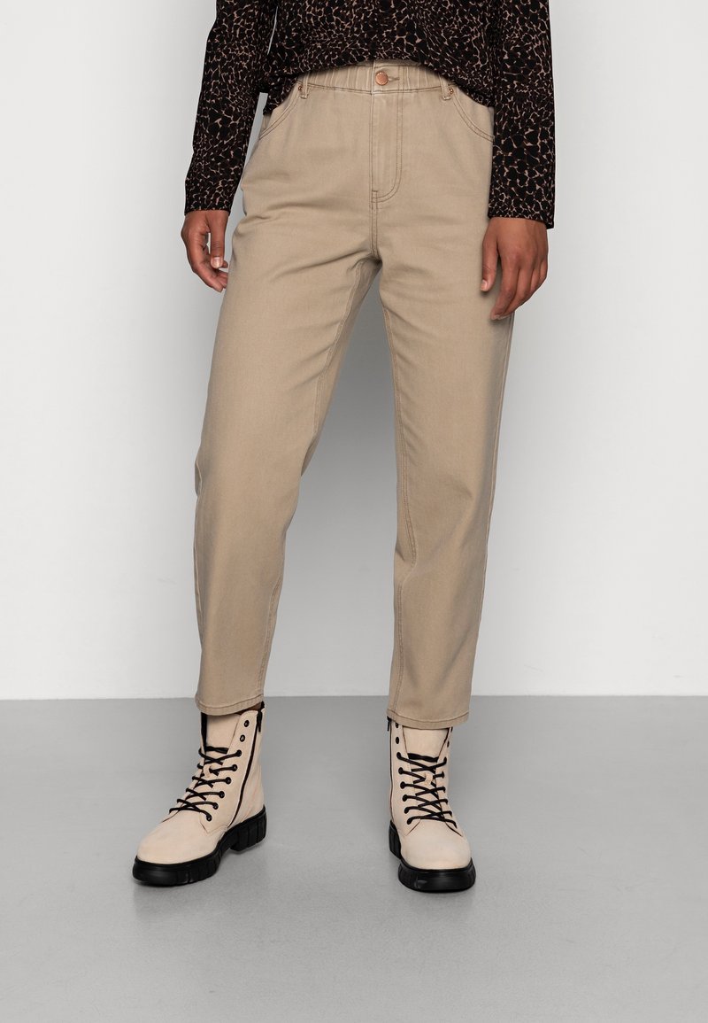 Los pantalones tapered color beige presentan una cintura alta y bolsillos frontales, combinados con botas color crema con cordones y suelas negras. El fondo es de un gris liso.