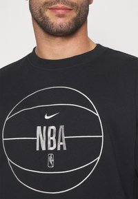 Homme portant un sweat-shirt noir avec un logo NBA de basket-ball blanc brodé et un swoosh Nike sur la poitrine.