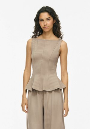 Frau mit lockigem dunklem Haar, die ein ärmelloses beiges gestreiftes Peplum-Top und passende weit geschnittene Hosen vor einem schlichten weißen Hintergrund trägt.