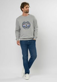 Lichtgrijs sweatshirt met een ronde halslijn en lange mouwen, voorzien van een geprinte logo in donkerblauw, wit en rood. Draagt de combinatie met blauwe jeans en witte sneakers.