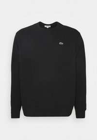 Sudadera negra con cuello redondo, mangas largas, puños de canalé y un pequeño logo verde de Lacoste en el pecho. Textura de tela suave.