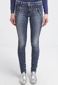 Jeans en denim skinny fit bleu foncé avec décoloration. Présentent des poches avant zippées et une texture lisse, associés à des chaussures argentées.
