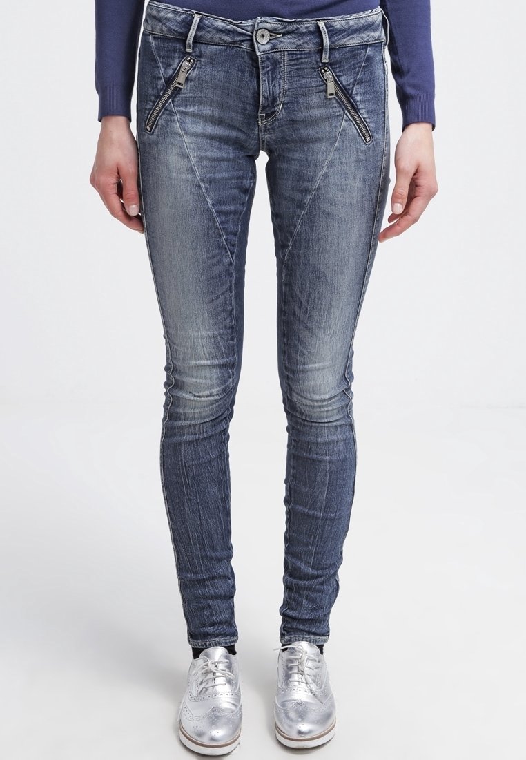 Jeans en denim skinny fit bleu foncé avec décoloration. Présentent des poches avant zippées et une texture lisse, associés à des chaussures argentées.