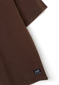 T-shirt in cotone marrone con maniche corte, caratterizzato da un collo rotondo e un'etichetta con testo bianco su uno sfondo scuro vicino all'orlo.