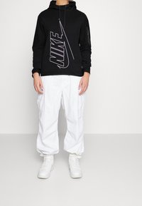 Sudadera negra con capucha y logo de Nike blanco, con tejido texturizado y cordón. Combinada con pantalones cargo blancos de corte holgado y zapatillas blancas.