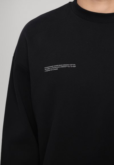 Schwarzer Pullover aus Bio-Baumwolle mit geripptem Kragen. Verfügt über weißen Text auf der Brust, der die Stoffbehandlung für Frische beschreibt.