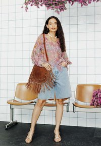 Blouse en dentelle florale, shorts en denim clair, sac en suède frangé marron avec des clous, et sandales dorées. L'arrière-plan comporte des bancs et des fleurs.