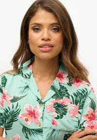 Camicia a maniche corte in tessuto turchese chiaro con un motivo floreale rosa e verde, colletto, e una piccola tasca frontale con dettagli di cucitura.