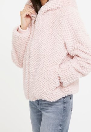 Kobieta ubrana w jasnoróżową, teksturowaną kurtkę z futerka ekologicznego z kapturem, zamkiem błyskawicznym i kieszeniami, w połączeniu z jasnoniebieskimi dżinsami.