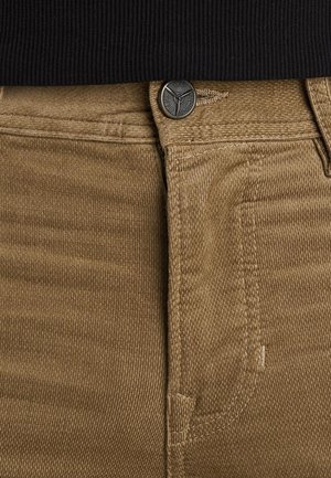 Beige ribfluweel stof met getextureerde ribbels, voorzien van een metalen knoopsluiting en gestikte details bij de zakken en de tailleband.