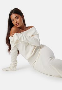 Top bianco a maglia con spalle scoperte e maniche lunghe, abbinato a una gonna aderente coordinata, che mette in evidenza una texture morbida e una silhouette slanciata.