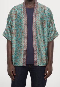 Kortærmet kimono-lignende jakke i turkis med indviklede blomster- og geometriske mønstre, der har en kontrasterende rød kant.