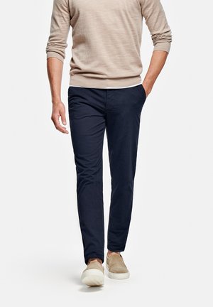 Marineblaues Slim-Fit-Hose, Baumwollmischung, mit Fronttaschen, kombiniert mit einem beigen Strickpullover und beigen Slippern, läuft vor einem schlichten Hintergrund.