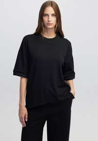 Černá oversized mikina s krátkým rukávem a kulatým výstřihem, vyrobená z hladkého materiálu, s volným střihem a bočními rozparky.