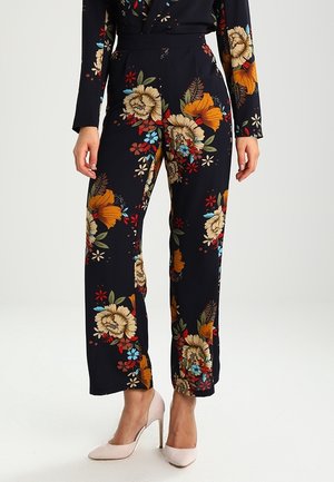Femme portant un pantalon noir avec de grands motifs floraux colorés, associé à des chaussures à talons hauts rose pâle, debout devant un fond uni.