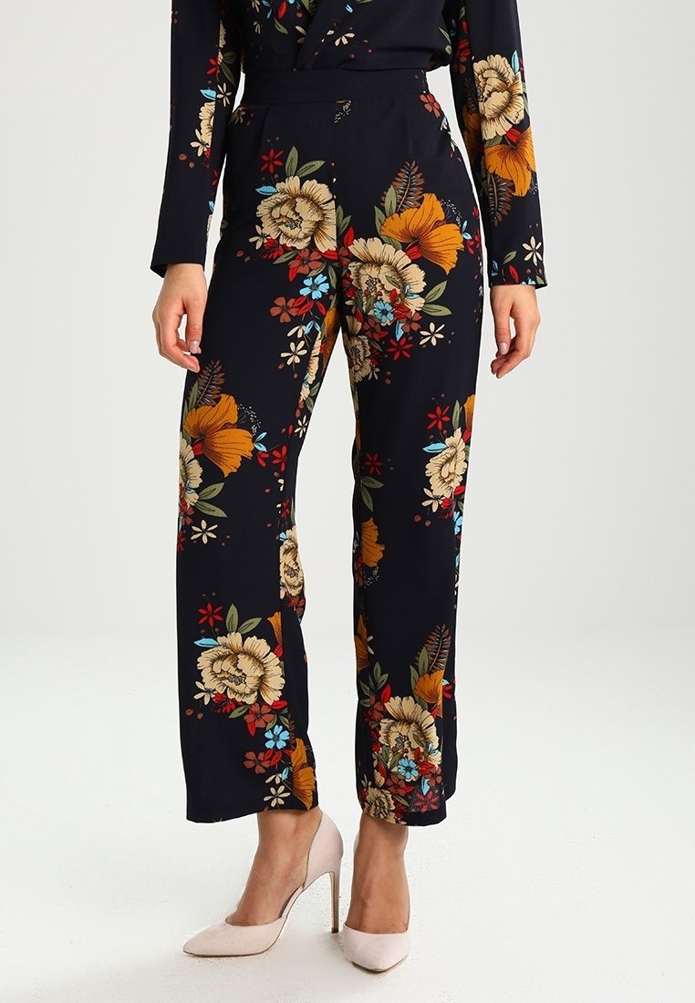 Femme portant un pantalon noir avec de grands motifs floraux colorés, associé à des chaussures à talons hauts rose pâle, debout devant un fond uni.