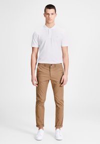 Weißes kurzärmliges Poloshirt mit Kragen, kombiniert mit braunen schmal geschnittenen Chinos und weißen Sneakern. Einfaches, sauberes Design mit subtiler Markenkennzeichnung.