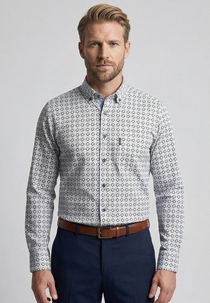 Homme aux cheveux blonds courts portant une chemise blanche à manches longues avec un motif géométrique bleu, ceinture marron et pantalon bleu marine, debout de manière neutre.