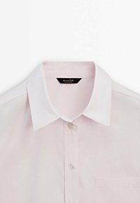 Chemise rose clair à boutons avec col blanc pointu, deux boutons visibles à l'avant et une poche poitrine du côté droit.