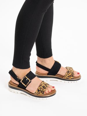 Füße mit schwarzen und Leopardenmuster-Sandalen mit dicken weißen Sohlen, kombiniert mit schwarzen, eng anliegenden Hosen.