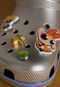 Sølv gummiklær med tre karakterformede charms, en grønn edelsten og en gyllen stjerneakent; teksturert overflate med ventilasjonsåpninger.