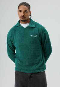 Groene fleece trui met een kraag en geborduurd logo "Essence of Life". Zachte textuur, losse pasvorm, gecombineerd met donkere spijkerbroek.