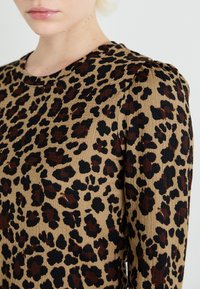 Top s leopardím vzorom z textúrovaného béžového materiálu, ktorý obsahuje čierne a hnedé škvrny. Golier je okrúhly a dizajn obsahuje dlhé rukávy.