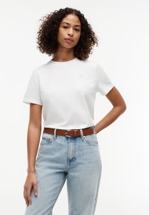 Frau mit lockigem Haar, die ein weißes T-Shirt trägt, das in hellblaue Jeans mit braunem Leder Gürtel gesteckt ist, posiert mit einer Hand in der Hüfte.