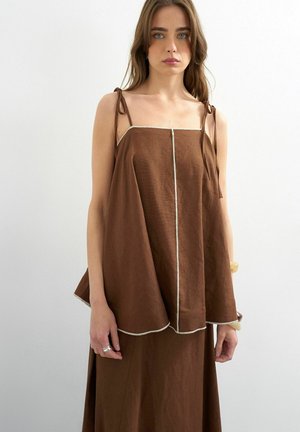 Vrouw met lang bruin haar draagt een losse, mouwloze bruine top met strikbandjes en een bijpassende rok, staand tegen een effen witte achtergrond.