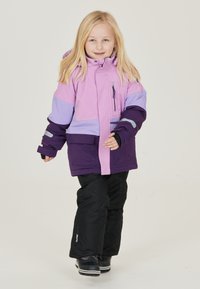 Lila und rosa isolierte Jacke mit Reißverschluss und Taschendetails, kombiniert mit schwarzen Hosen und Schuhen, entworfen für den Einsatz bei kaltem Wetter.