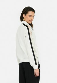 Sweatshirt blanc avec des rayures noires sur les côtés et des détails en dentelle le long des manches. Présente un col haut et des poignets côtelés pour une texture.