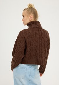 Brown cable-knit sweater with a high collar and oversized fit, featuring ribbed cuffs and hem. Worn over a light blue denim skirt.