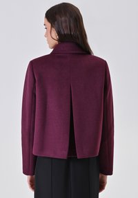 Giacca corta bordeaux con collo alto e dettaglio con piega posteriore, realizzata in tessuto strutturato. Design senza cuciture con maniche lunghe.