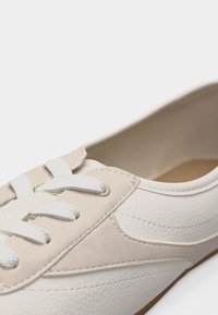 Κοντινό πλάνο ενός λευκού και μπεζ αθλητικού παπουτσιού που δείχνει τα κορδόνια, το υλικό της επάνω επιφάνειας με υφή και τις λεπτομέρειες ραφής σε απλό φόντο.