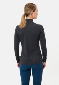 Frau mit blonden Pferdeschwanz, die ein langärmliges schwarzes Sportoberteil und eine blaue Hose trägt, steht mit dem Rücken zu einem weißen Hintergrund.