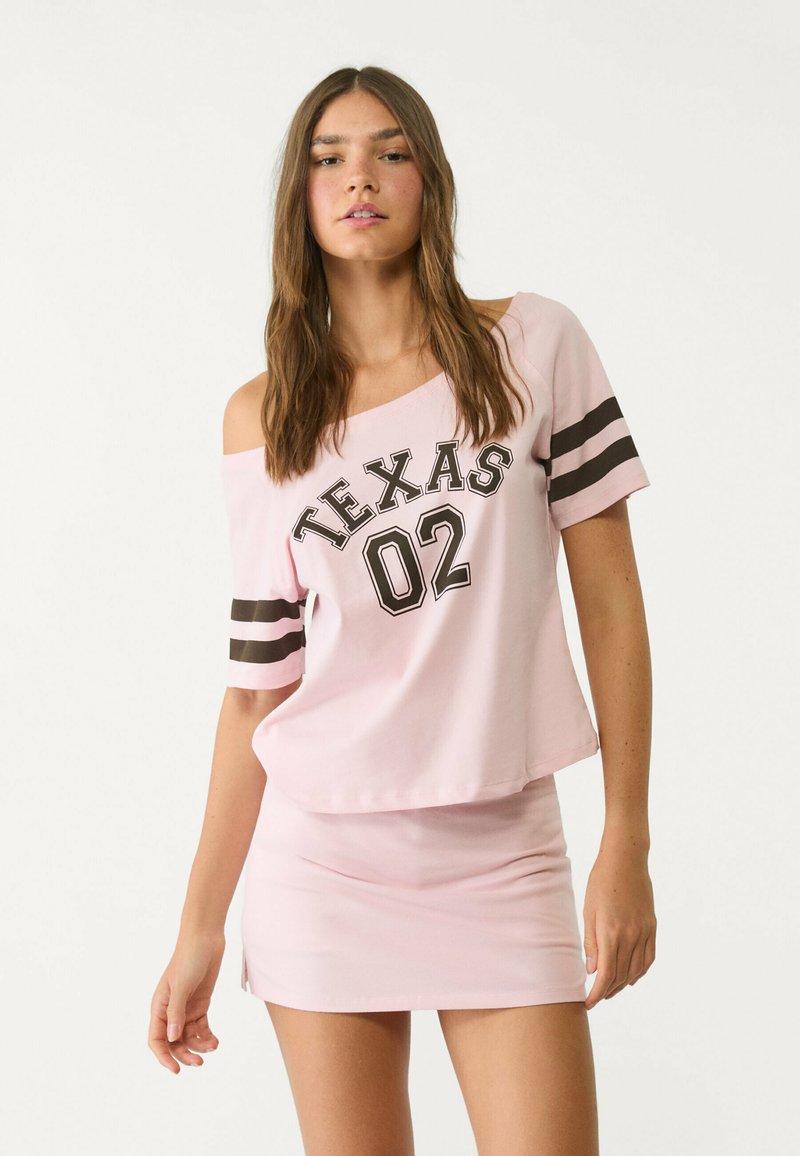 Roze off-shoulder shirt met "TEXAS 02" graphic, korte mouwen met zwarte strepen, gecombineerd met een bijpassende roze mini rok. Gladde stof.