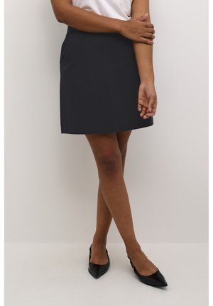 Jupe mini bleue marine en tissu lisse, coupe droite, avec des coutures discrètes. Associée à des ballerines noires à bout pointu.