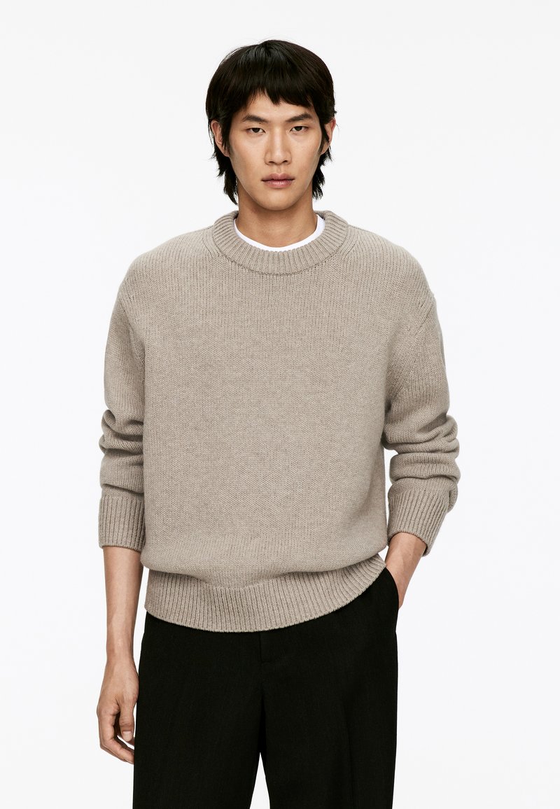 Helle beige gestrickte Sweater mit rundem Ausschnitt und gerippten Bündchen und Saum. Über einem weißen Hemd getragen, kombiniert mit dunklen Hosen.