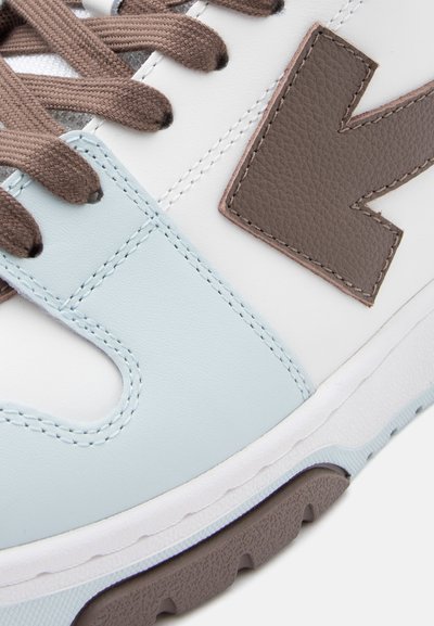 Εικόνα στενής λήψης ενός αθλητικού παπουτσιού με καφέ κορδόνια, λευκές και ανοιχτομπλε δερμάτινες επιφάνειες, μεγάλο καφέ υφασμένο λογότυπο "V" και υφασμένη καφέ σόλα.