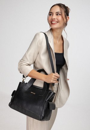 Glunderende vrouw in beige pak draagt een grote zwarte leren handtas met schouderband, kijkt zijwaarts tegen een effen achtergrond.